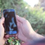 Mãos seguram um smartphone Samsung ao ar livre, com vegetação desfocada ao fundo.