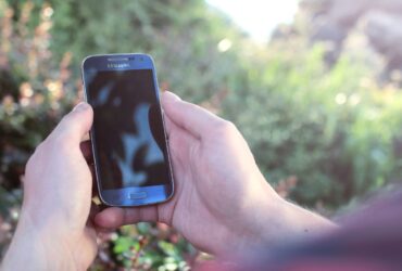 Mãos seguram um smartphone Samsung ao ar livre, com vegetação desfocada ao fundo.