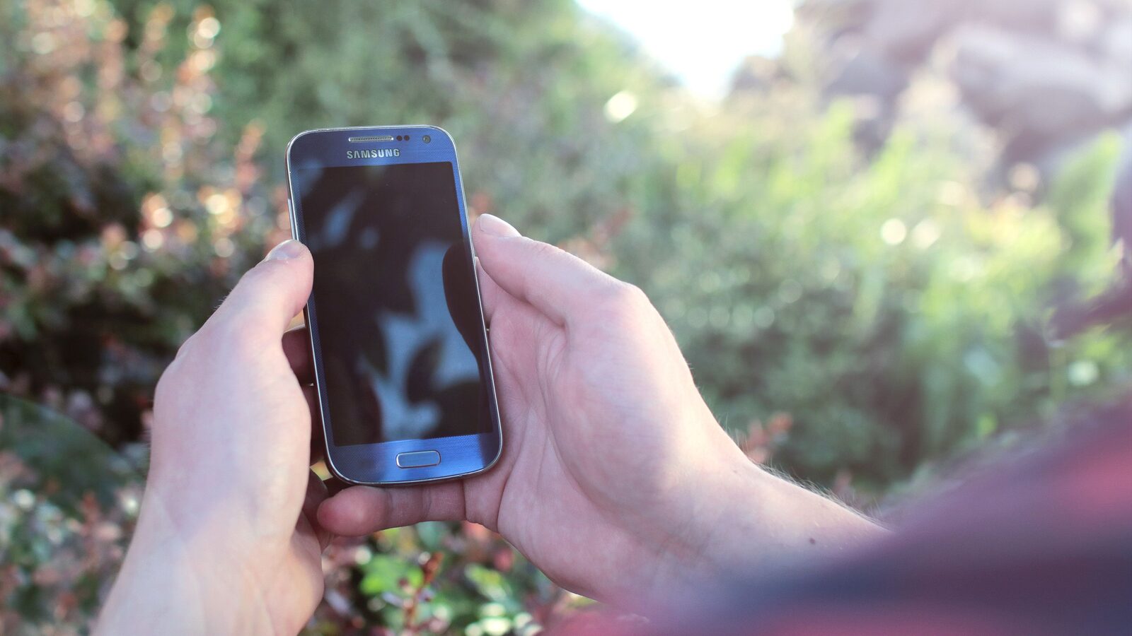 Mãos seguram um smartphone Samsung ao ar livre, com vegetação desfocada ao fundo.