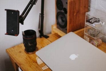 MacBook fechado sobre uma secretária de madeira, junto a equipamento de fotografia e som.