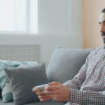 Homem sentado num sofá usa auscultadores e segura um telemóvel numa sala iluminada.