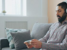 Homem sentado num sofá usa auscultadores e segura um telemóvel numa sala iluminada.
