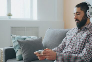 Homem sentado num sofá usa auscultadores e segura um telemóvel numa sala iluminada.