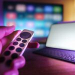 Mão segura um comando diante de um televisor desfocado e de um tablet com teclado sobre a mesa.