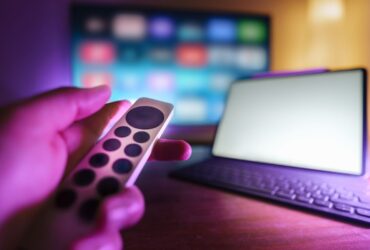 Mão segura um comando diante de um televisor desfocado e de um tablet com teclado sobre a mesa.