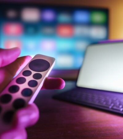 Mão segura um comando diante de um televisor desfocado e de um tablet com teclado sobre a mesa.