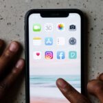 Mãos junto de um smartphone com ícones de aplicações, incluindo Facebook e Instagram, visíveis no ecrã.