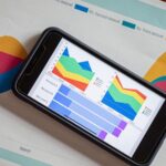 Smartphone sobre folhas com gráficos coloridos, junto a dois lápis e uma lupa.