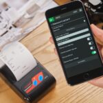 Mão segura um smartphone ao lado de uma impressora que exibe um recibo de papel sobre uma mesa de madeira.