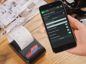 Mão segura um smartphone ao lado de uma impressora que exibe um recibo de papel sobre uma mesa de madeira.