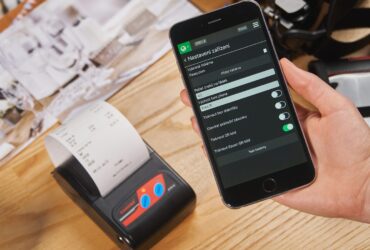 Mão segura um smartphone ao lado de uma impressora que exibe um recibo de papel sobre uma mesa de madeira.