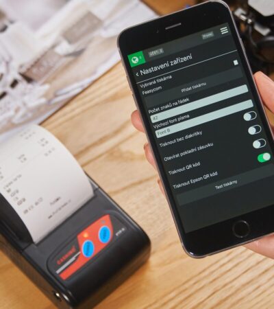 Mão segura um smartphone ao lado de uma impressora que exibe um recibo de papel sobre uma mesa de madeira.