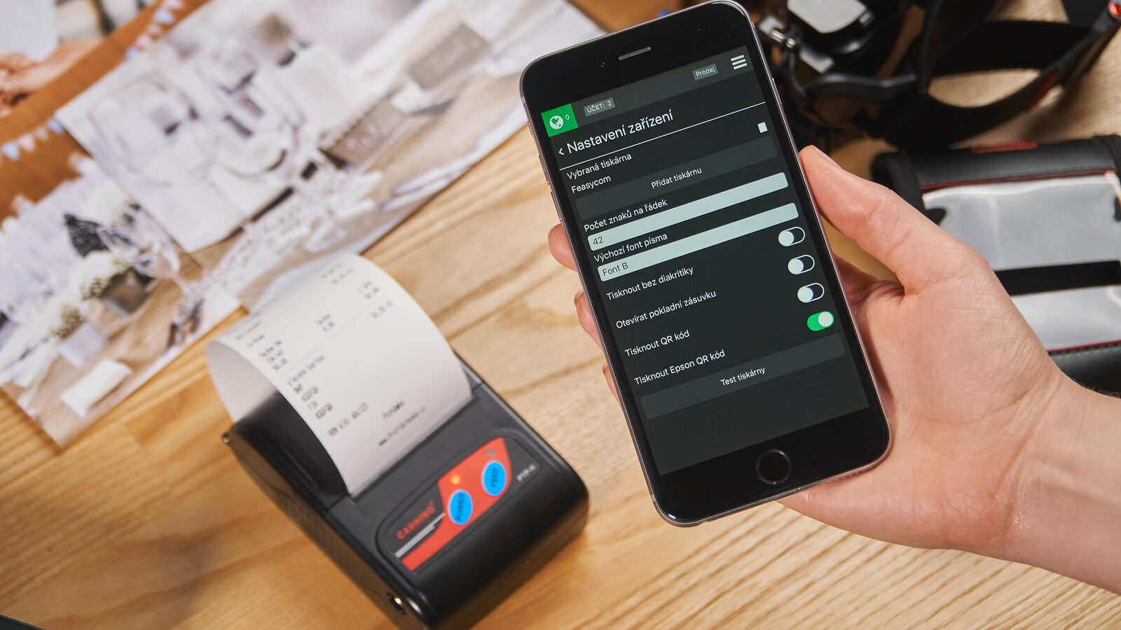 Mão segura um smartphone ao lado de uma impressora que exibe um recibo de papel sobre uma mesa de madeira.