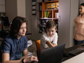 Dois estudantes usam computadores em uma sala de aula enquanto uma professora observa.