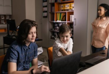 Dois estudantes usam computadores em uma sala de aula enquanto uma professora observa.