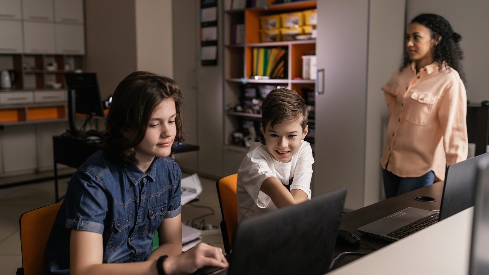 Dois estudantes usam computadores em uma sala de aula enquanto uma professora observa.
