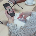Pessoa segura um telemóvel junto a microfones e a um bloco de notas preparado para gravar um podcast.