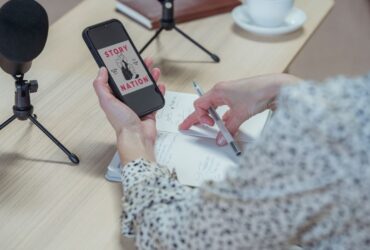 Pessoa segura um telemóvel junto a microfones e a um bloco de notas preparado para gravar um podcast.