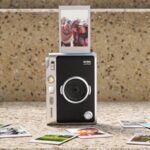 Câmara Instax Mini Evo 2 com uma fotografia a sair e várias impressões instantâneas dispostas à frente.
