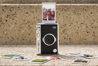 Câmara Instax Mini Evo 2 com uma fotografia a sair e várias impressões instantâneas dispostas à frente.