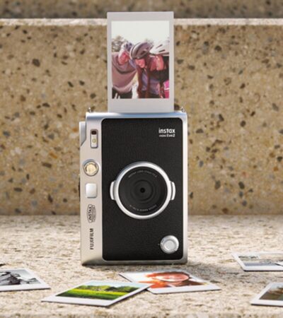Câmara Instax Mini Evo 2 com uma fotografia a sair e várias impressões instantâneas dispostas à frente.