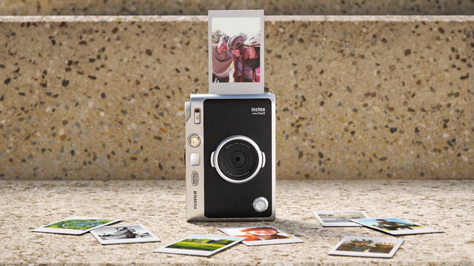Câmara Instax Mini Evo 2 com uma fotografia a sair e várias impressões instantâneas dispostas à frente.