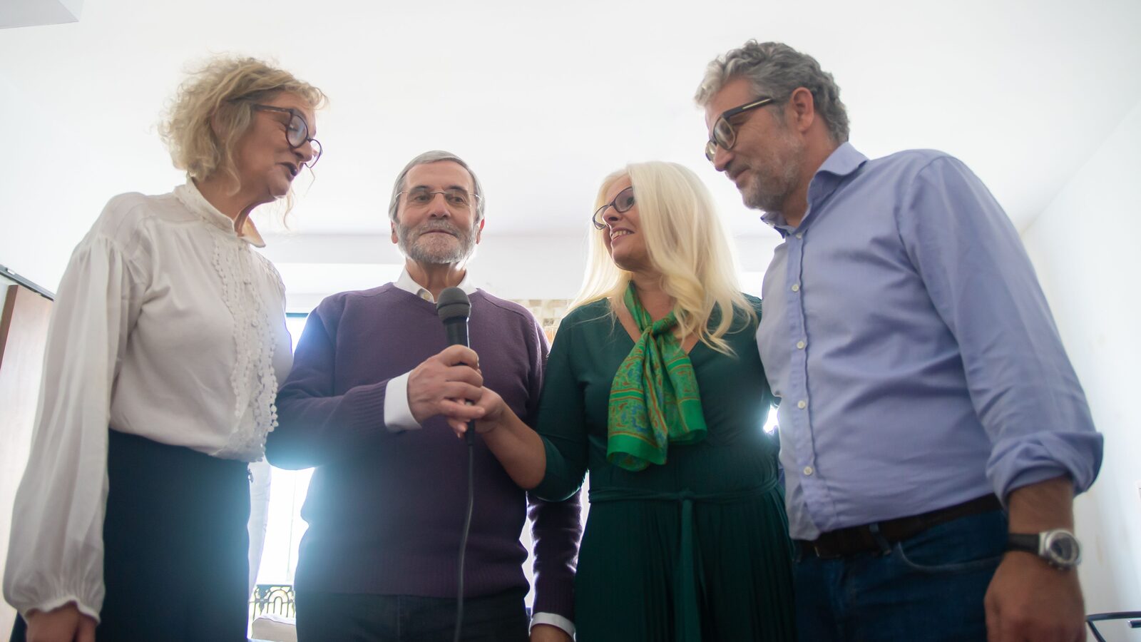Quatro adultos participam numa sessão de karaoke com um microfone ao centro.
