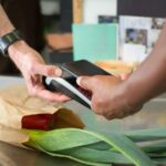 Pessoa aproxima um smartphone de um terminal de pagamento numa caixa de supermercado.
