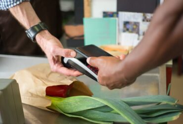 Pessoa aproxima um smartphone de um terminal de pagamento numa caixa de supermercado.