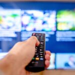 Mão aponta um comando para um televisor com o ecrã desfocado