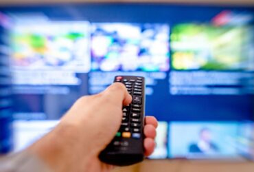 Mão aponta um comando para um televisor com o ecrã desfocado