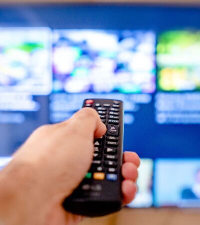 Mão aponta um comando para um televisor com o ecrã desfocado