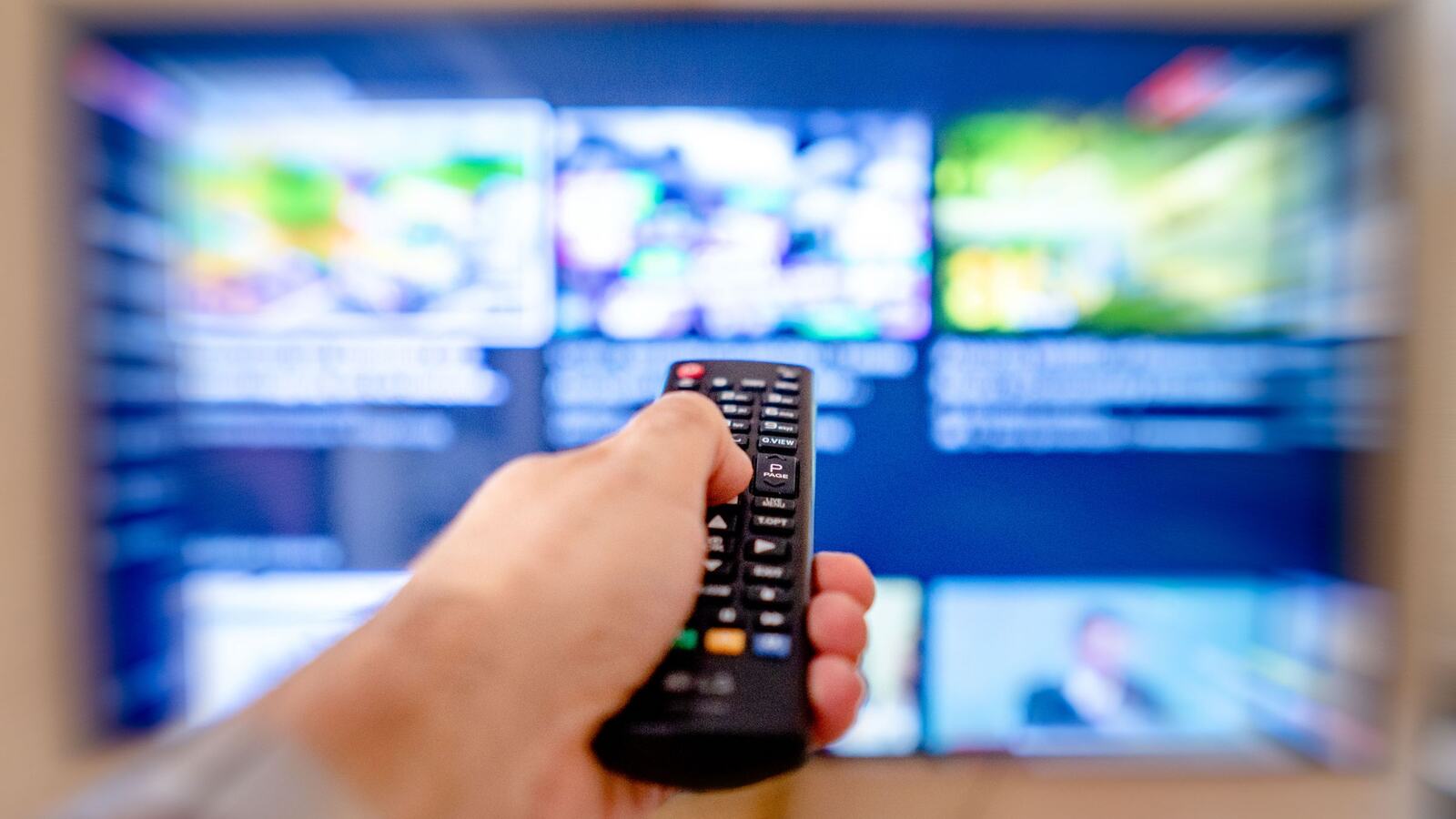 Mão aponta um comando para um televisor com o ecrã desfocado
