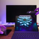 Mão aponta um comando para um televisor, com um tablet e um telemóvel sobre a mesa.