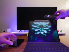 Mão aponta um comando para um televisor, com um tablet e um telemóvel sobre a mesa.