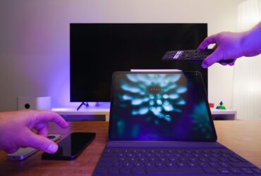 Mão aponta um comando para um televisor, com um tablet e um telemóvel sobre a mesa.