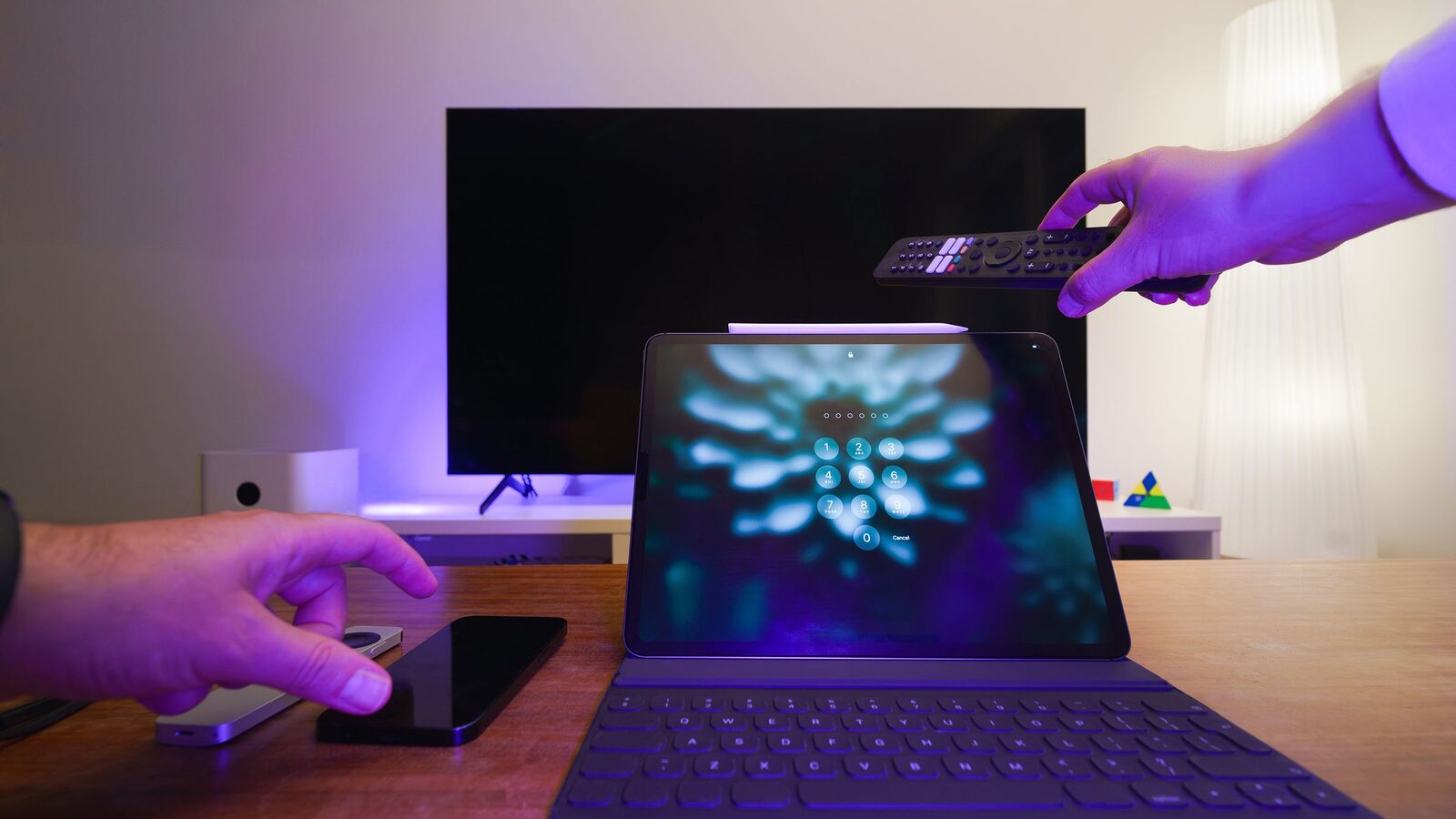 Mão aponta um comando para um televisor, com um tablet e um telemóvel sobre a mesa.