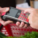 Mãos introduzem um PIN em um terminal de pagamento junto a uma cesta de compras.