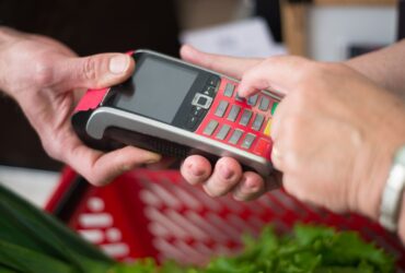 Mãos introduzem um PIN em um terminal de pagamento junto a uma cesta de compras.