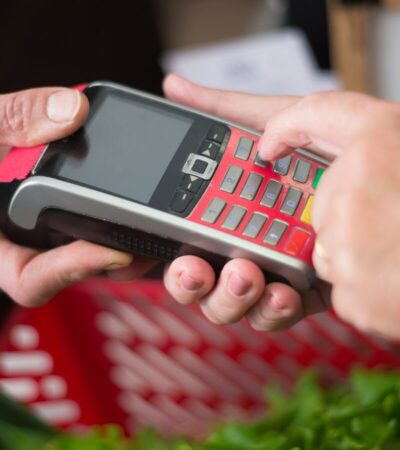 Mãos introduzem um PIN em um terminal de pagamento junto a uma cesta de compras.