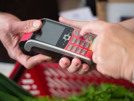 Mãos introduzem um PIN em um terminal de pagamento junto a uma cesta de compras.