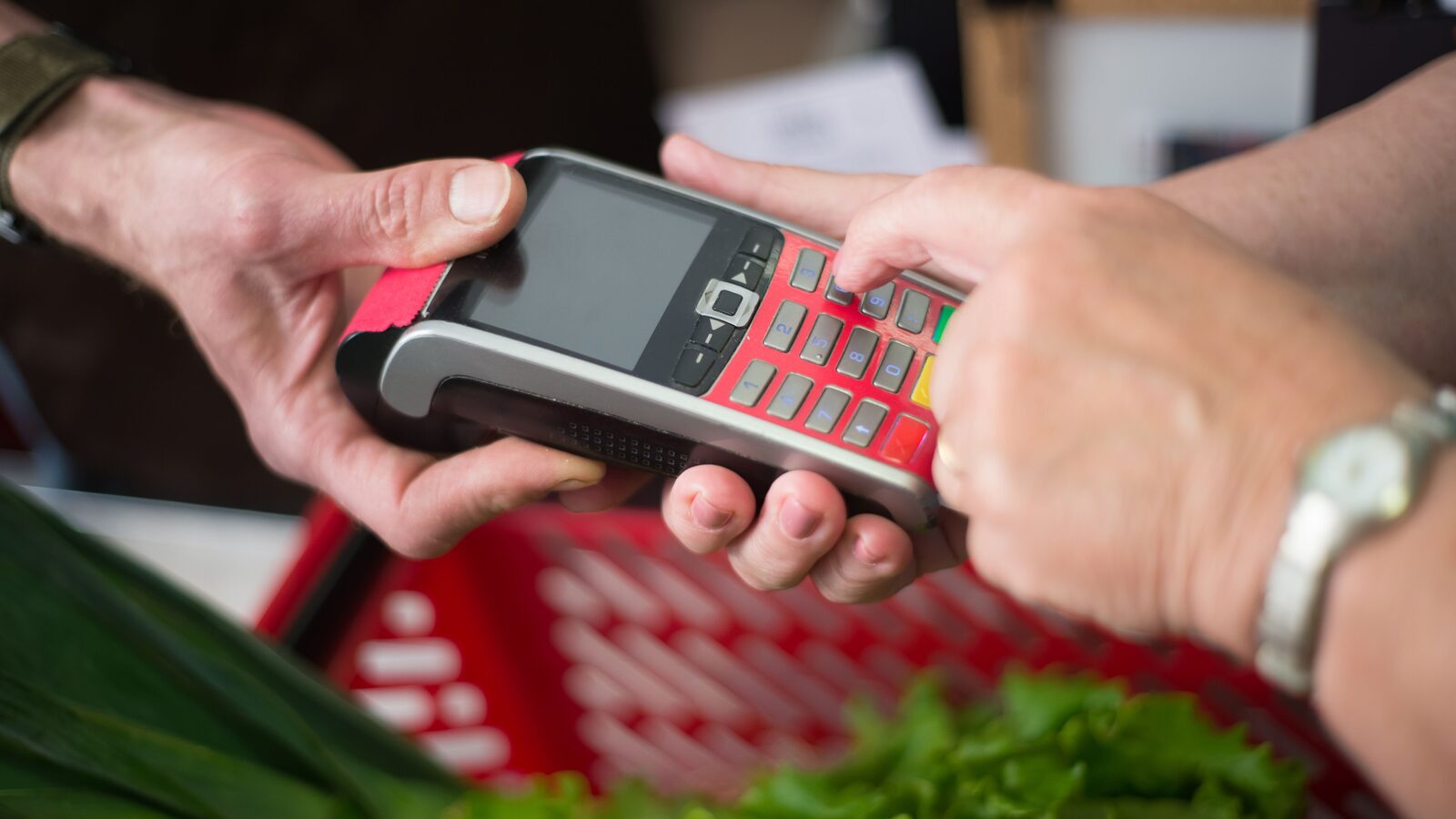 Mãos introduzem um PIN em um terminal de pagamento junto a uma cesta de compras.
