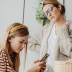Mãe observa uma adolescente que usa um smartphone à mesa.