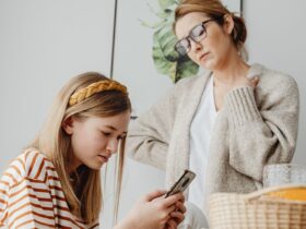 Mãe observa uma adolescente que usa um smartphone à mesa.