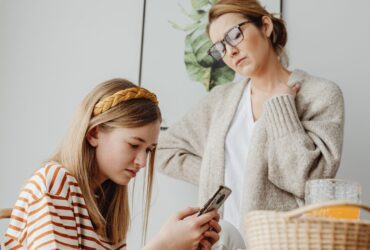 Mãe observa uma adolescente que usa um smartphone à mesa.