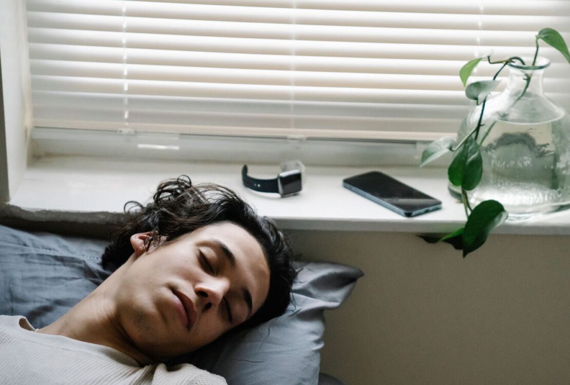 Pessoa a dormir junto a uma janela, com um smartwatch e um smartphone pousados no parapeito.