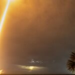 Rasto luminoso de um Falcon 9 com satélites Starlink durante um lançamento noturno, visto sobre a água e entre palmeiras.