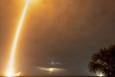 Rasto luminoso de um Falcon 9 com satélites Starlink durante um lançamento noturno, visto sobre a água e entre palmeiras.