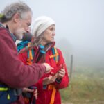 Dois caminhantes com mochilas consultam um telemóvel numa paisagem rural com nevoeiro.
