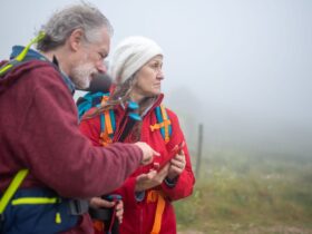 Dois caminhantes com mochilas consultam um telemóvel numa paisagem rural com nevoeiro.